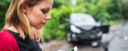 A close-up of a young woman with smartphone by the damaged car after a car accident, text messaging. Copy space.