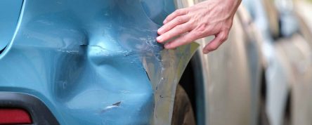 Driver hand examining dented car with damaged fender parked on city street side. Road safety and vehicle insurance concept.